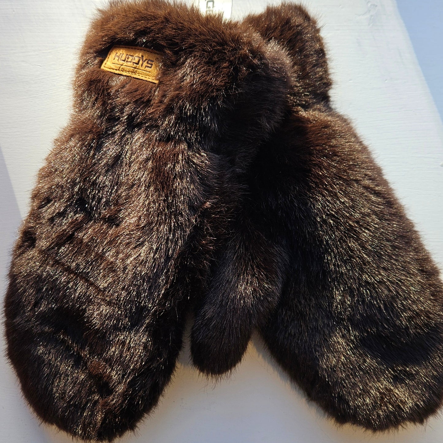Pair of brown fur mittens on a white surface with a visible brand tag.