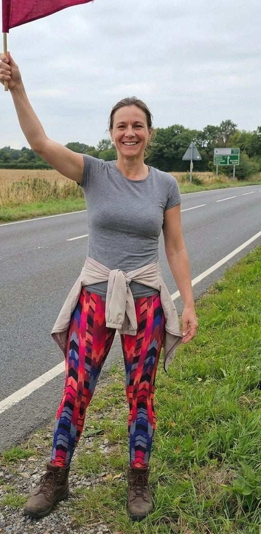 Woman standing on the side of a road holding a red flag, wearing a gray t-shirt and colorful leggings.