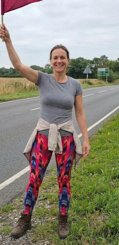 Woman standing on the side of a road holding a red flag, wearing a gray t-shirt and colorful leggings.